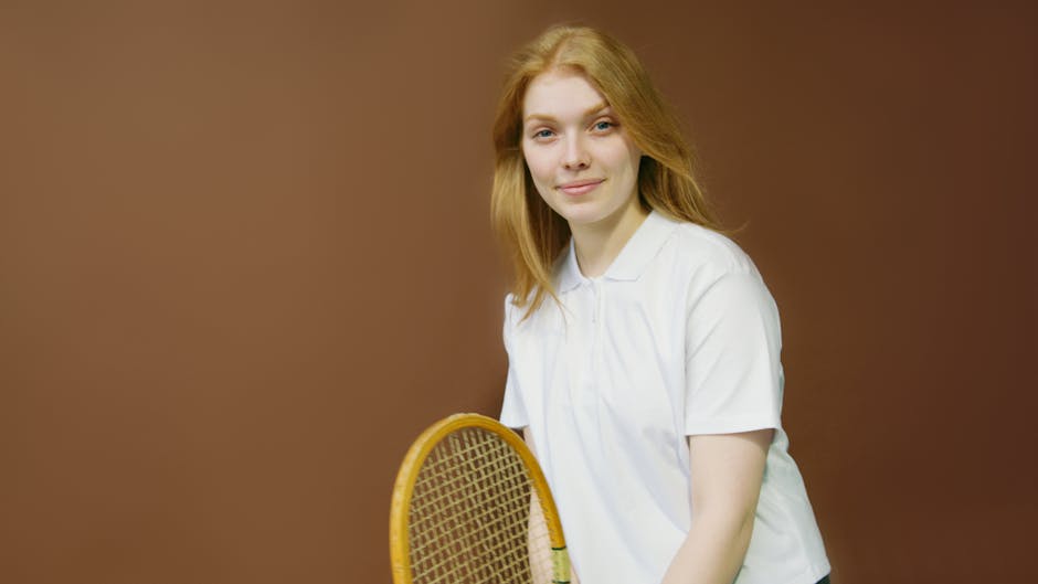 A smiling woman holds a tennis racket indoors, wearing a white shirt, ready for an athletic activity.