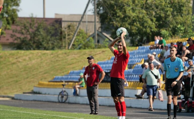 Soccer player prepares to throw in the ball during an outdoor match, with referee and spectators watching.