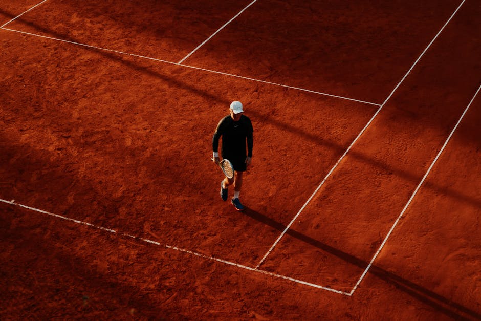 An overhead shot of a tennis player on a clay court during the day, with long shadows.