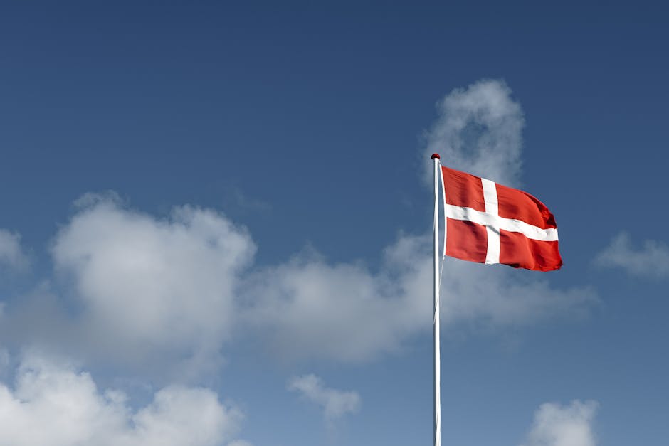 A beautiful view of the Danish flag waving high against a vivid blue sky with fluffy clouds.