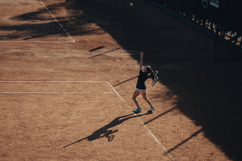 Aerial view of a woman playing tennis, serving on a clay court under sunlight.