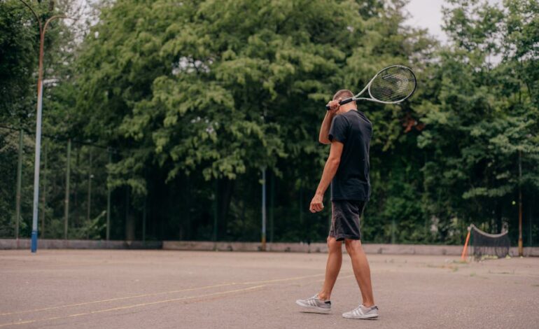 A young man holds a tennis racket while standing on an outdoor court surrounded by trees.