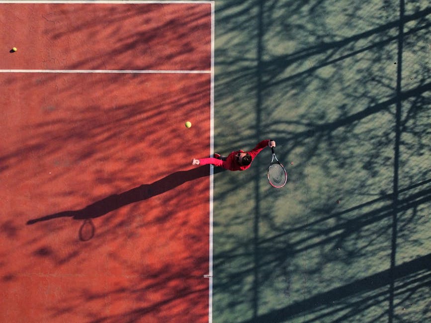 Aerial view of a tennis player serving on a colorful outdoor court with long shadows.