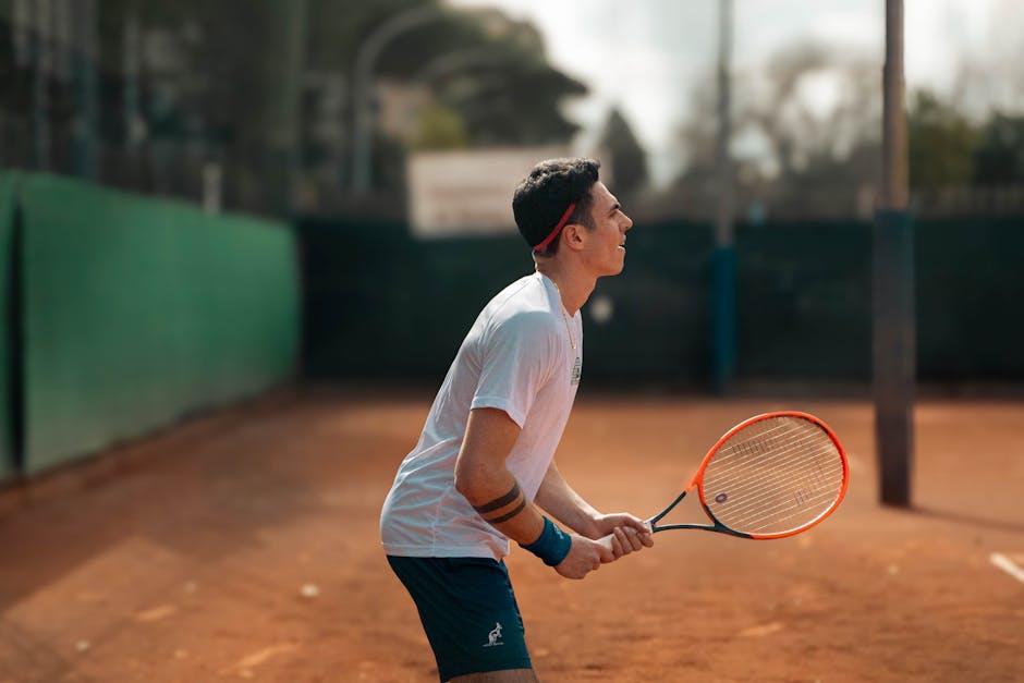 A young man in sportswear holding a racket, ready to serve on a sunny clay tennis court.