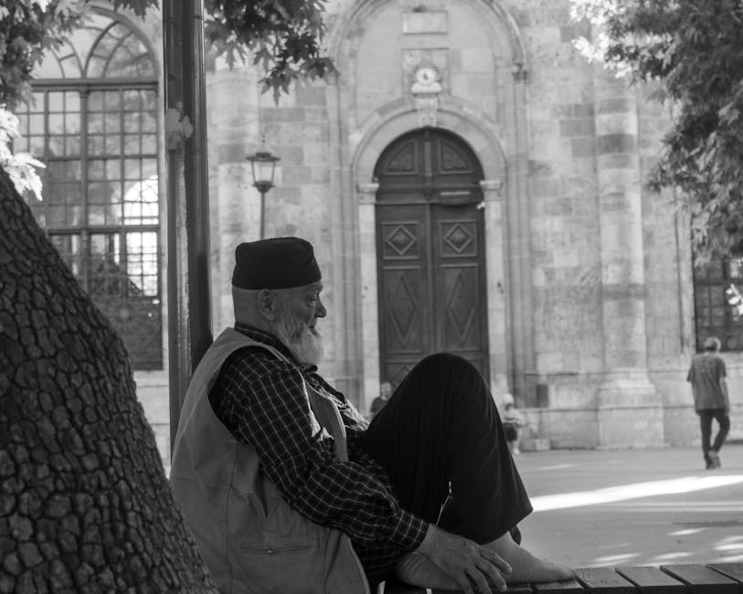 A serene monochrome image of an elderly man sitting on a bench near an ornate building facade.