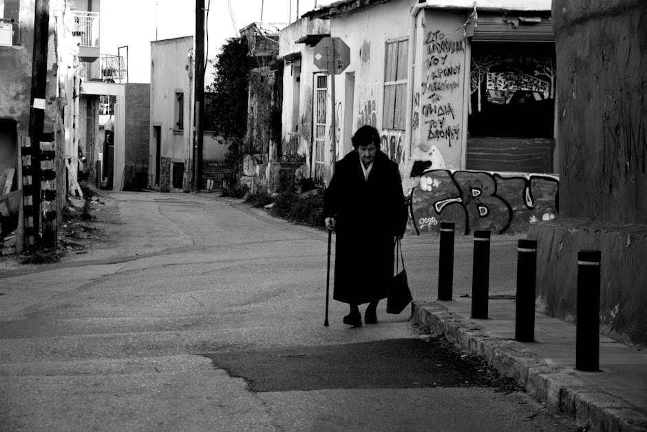 Black and white image of an elderly woman walking with a cane on a graffiti-lined urban street.