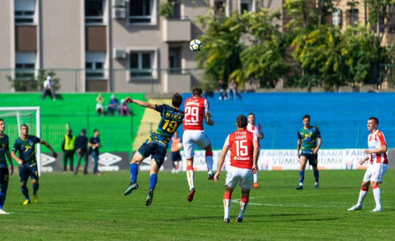 Soccer players in red and blue uniforms compete fiercely during an outdoor match on a sunny day.