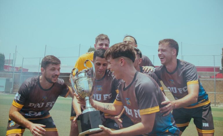 Soccer team joyfully celebrating their victory on a sunny pitch holding a trophy.