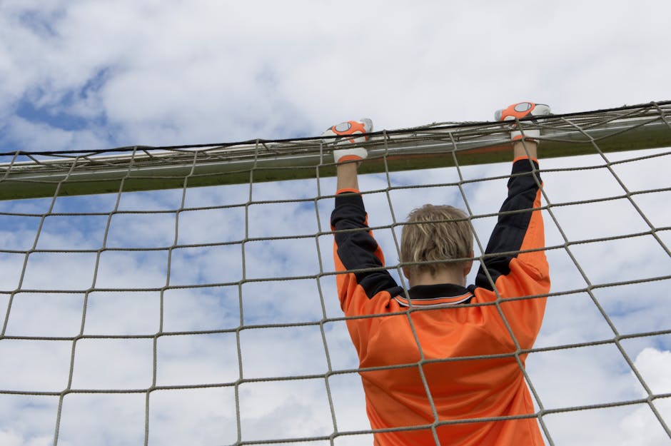 A young soccer goalie in orange kit hangs from the goal post on a cloudy day.