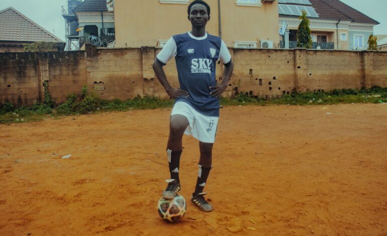 A teenage soccer player in uniform poses with a ball on a dirt field.