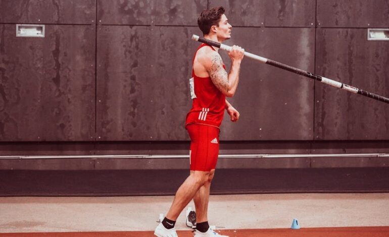 Athletic male in sportswear holding pole for pole vault indoors, showing determination and focus.