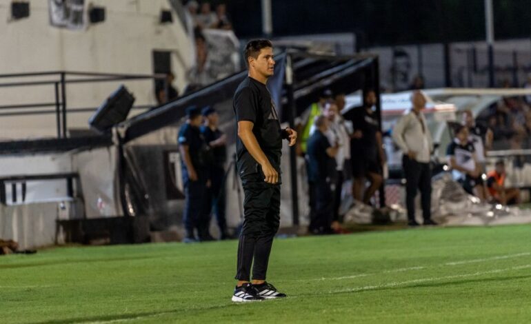 Focused football coach stands on the field sidelines during an intense match at night.