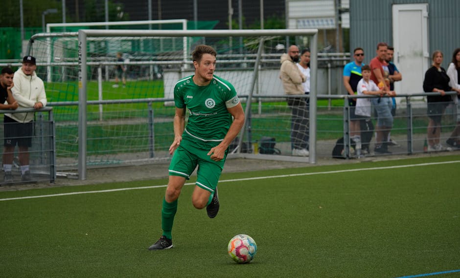 Adult male soccer player in green jersey dribbling ball on pitch during daytime match with spectators.