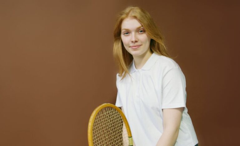 A smiling woman holds a tennis racket indoors, wearing a white shirt, ready for an athletic activity.