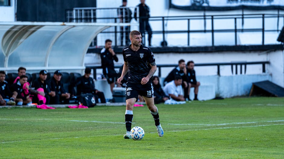 Soccer player dribbling during a match with team watching from the sidelines.