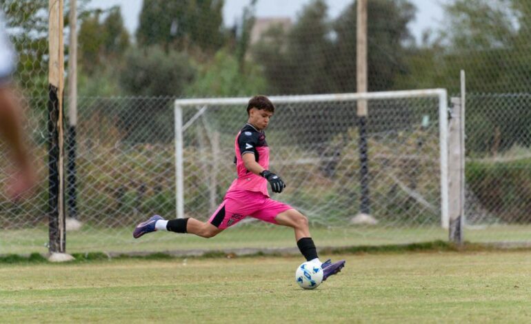 A young goalkeeper in pink uniform kicks a soccer ball on an outdoor field.