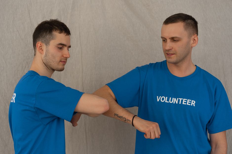 Two male volunteers in blue shirts exchange an elbow bump, symbolizing support and teamwork.