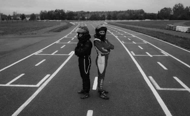 Two karting racers standing back-to-back on a racetrack in full gear, showcasing determination in monochrome.