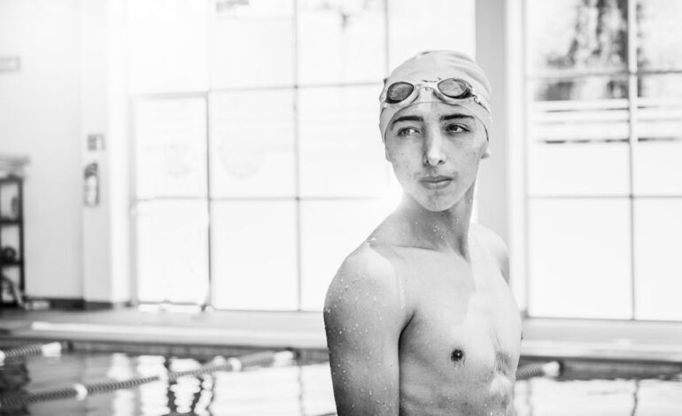 Monochrome image of a shirtless teenage swimmer with goggles in an indoor pool.