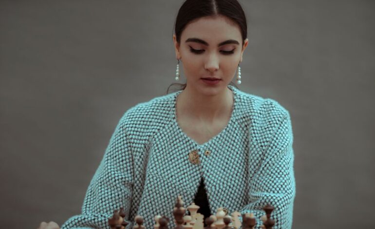 Concentrated ethnic female player sitting at table and playing chess board game of strategic skill against gray background