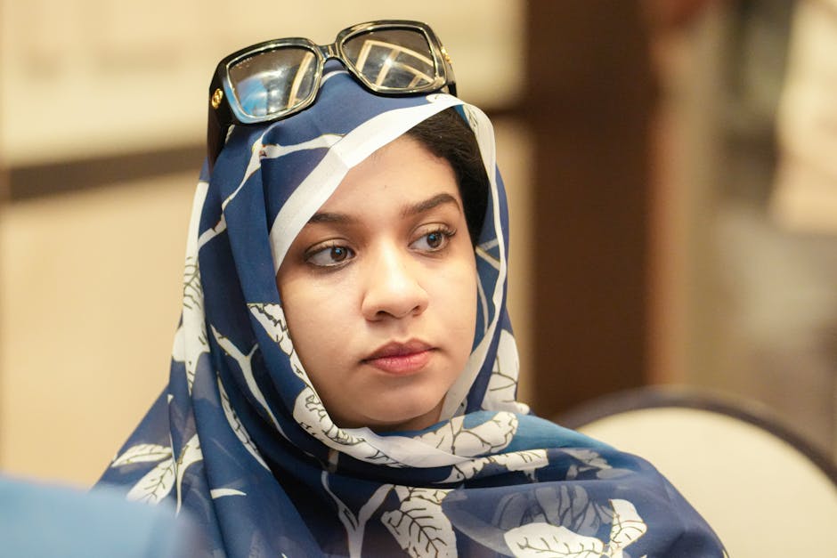 Portrait of an adult woman wearing a blue floral hijab with sunglasses on her head, showcasing thoughtful expression indoors.