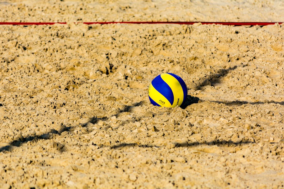 A vibrant yellow and blue volleyball resting on the sand at a beach with clear afternoon sunlight.