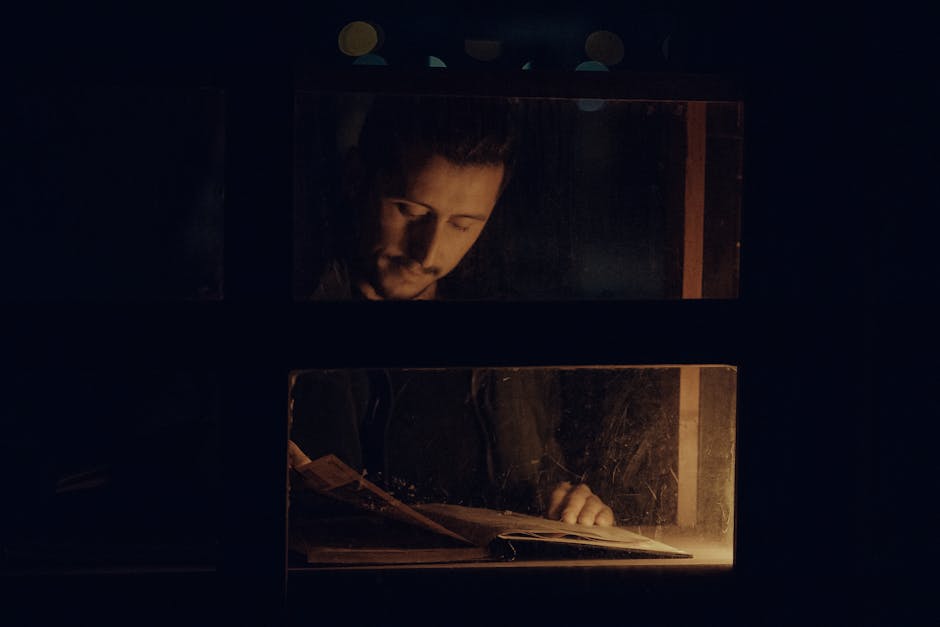 A contemplative man reading a book indoors by dim light, seen through a window at night.