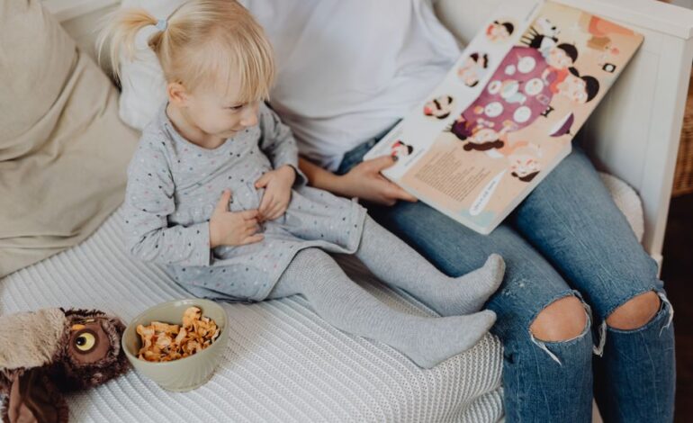 Mother reading a book to her daughter in a cozy bedroom setting with snacks nearby.