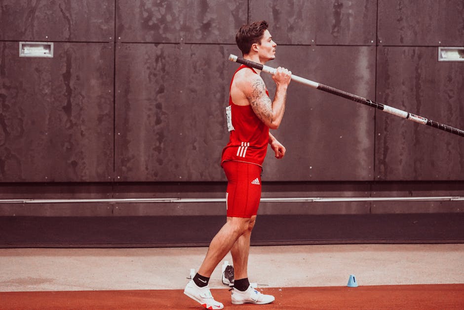 Athletic male in sportswear holding pole for pole vault indoors, showing determination and focus.