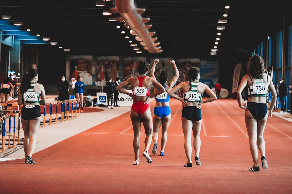 Group of female athletes at the starting line of an indoor track race, wearing numbered sportswear.