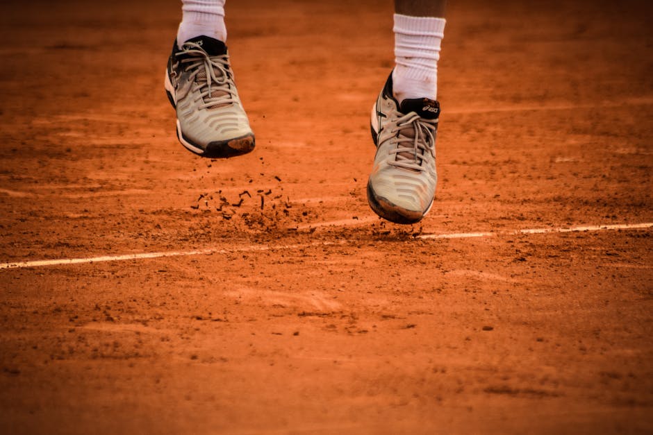 Close-up of an athlete's shoes mid-jump on a clay tennis court.