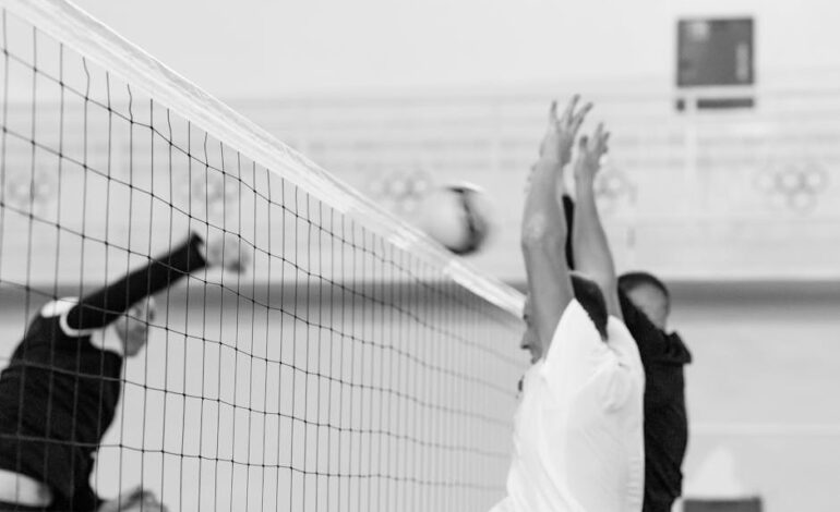 Black and white photo capturing players in mid-air blocking shot at volleyball net.
