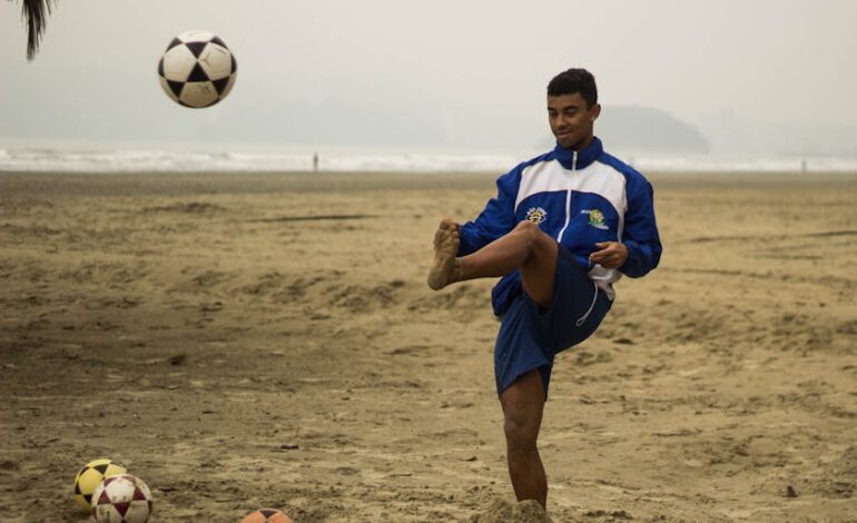 A young male athlete practices soccer on a sandy beach, showcasing skill under cloudy skies.