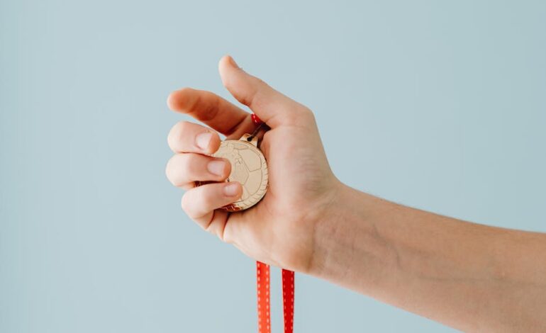 A close-up image of a hand holding a gold medal with a red ribbon against a blue background.